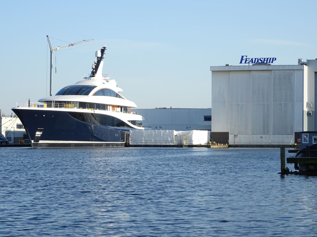 A dark-hulled yacht moored outside the white Feadship construction hall in Aalsmeer
