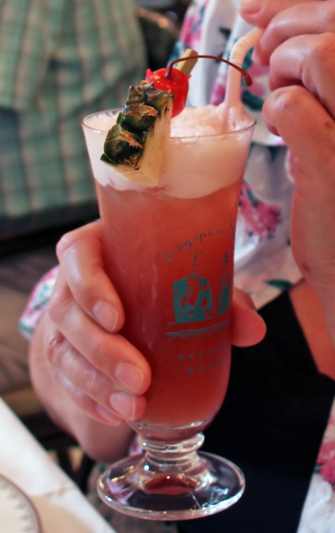 A pink Singapore Sling cocktail garnished with pineapple and a cherry, held up at Raffles
